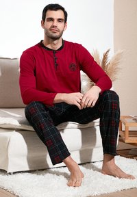 Rød langærmet top med rund hals og sorte knapper, parret med sorte ternede bukser med røde accenter, på en lysfarvet sofa.