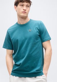 Τιρκουάζ βαμβακερό μπλουζάκι με στρογγυλή λαιμόκοψη, κοντά μανίκια και ένα μικρό λογότυπο στο αριστερό στήθος. Λεπτό ύφασμα, άνετη εφαρμογή.