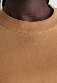 Brun ribbet T-skjorte med rund hals. Stoffet ser mykt og elastisk ut, med synlige sømmer langs kragen.