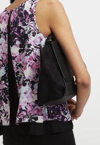 Černá kabelka s hladkou koženou texturou a jemným krokodýlím vzorem, s pozlaceným kováním. Nositelná na rameni s květinovým topem.