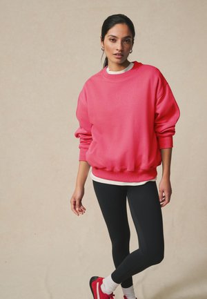 Kvinde iført en løs, lyserød sweatshirt, sorte leggings, hvide sokker og røde sneakers står mod en ensfarvet beige baggrund.