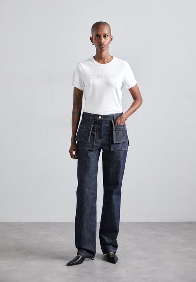 Valkoinen t-paita, jossa on kohokuvioitu "HELMUT LANG" -logo, yhdistettynä tummiin farkkuihin, joissa on kaksi suurta etureppua ja suora lahje.