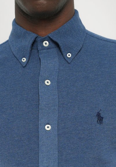 Chemise à boutons dans un tissu bleu texturé avec un col pointe. Elle présente des boutons blancs avec des coutures foncées et un petit logo brodé.