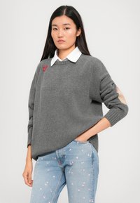 Gray knitted sweater with ribbed hem, featuring a red heart embroidery and light-colored elbow patches, worn over a white collared shirt.