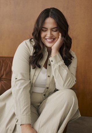 Chemise à rayures, boutonnée, beige et blanche, associée à un pantalon assorti. Texture lisse avec manches retroussées, boutons marron.