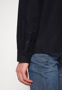 Svart bomullslångärmad skjorta med knäppta manschetter, med en texturerad tyg och en lätt lyster, ihop med blå denimjeans.