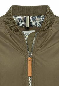 Olijfgroene bomberjack met een geribde kraag, ritssluiting en een oranje ritsaanhanger. De binnenvoering heeft een camouflagepatroon.