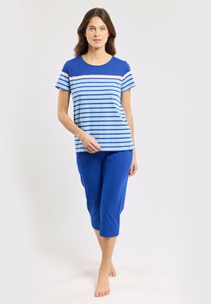 Femme marchant pieds nus portant un t-shirt à manches courtes rayé bleu et bleu clair et un pantalon capri bleu uni sur un fond uni.