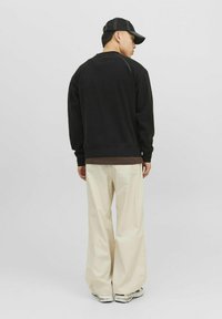 Čierna mikina s okrúhlym výstrihom a rebrovaným lemom, v kombinácii s voľnými béžovými nohavicami. Model má na sebe čiernu čiapku s bielym prešitím.