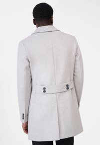 Cappotto grigio in lana con colletto ampio, cintura posteriore doppiopetto e tessuto texturizzato. Vestibilità su misura con tasche laterali e dettagli a bottoni.