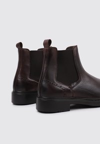 Bottines Chelsea en cuir marron avec des panneaux latéraux en élastique, bout rond et une semelle en caoutchouc noir bas. Présente des coutures décoratives sur le dessus.