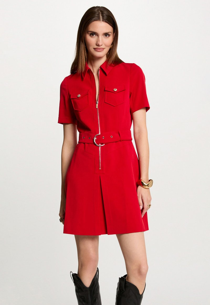 Vestido rojo de manga corta con cremallera frontal, dos bolsillos en el pecho, cintura ceñida con hebilla redonda y detalle de falda plisada. Tejido texturizado.