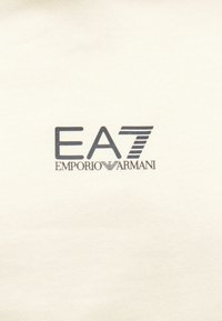 Kremowa tkanina ozdobiona logo EA7 i Emporio Armani w ciemnoniebieskim kolorze, z cyfrą "7" zaakcentowaną paskami. Gładka tekstura, minimalistyczny design.