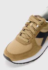 Beige sneaker featuring a mesh upper, suede accents, black logo, and flat laces. Rubber outsole with white midsole for cushioning.