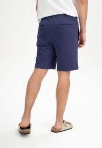 Marineblaue Baumwollshorts mit einem elastischen Bund, zwei Gesäßtaschen und einem lockeren Schnitt, kombiniert mit helltanen Slippern.