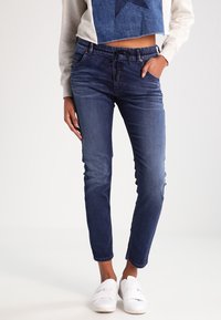 Vaqueros de mezclilla azul oscuro con pernera ajustada, cinturilla elástica y bolsillos frontales. Combinados con un suéter corto gris y zapatillas blancas sin cordones.