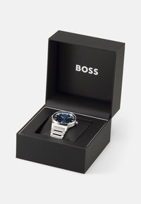 Silver wristwatch with a blue dial, located in a black box. Features a stainless steel band and a minimalist design, showcasing the brand name "BOSS."