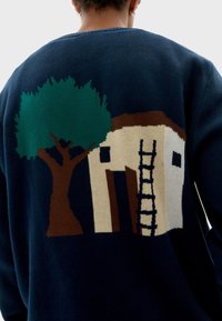 Pull en tricot marine avec un graphique d'un arbre brun et d'une maison avec une échelle en vert, beige et noir. Texture douce, coupe décontractée.