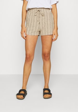 Vrouw draagt beige gestreepte shorts met trekkoord, wit ingestopt shirt en zwarte sandalen met twee bandjes, staand tegen een witte achtergrond.