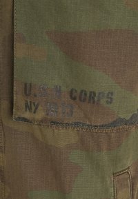 Vojenská kamuflážní tkanina v odstínech zelené a hnědé, s potiskem textu "U.S.N CORPS NY 1913" v tučné černé barvě. Texturovaný materiál.