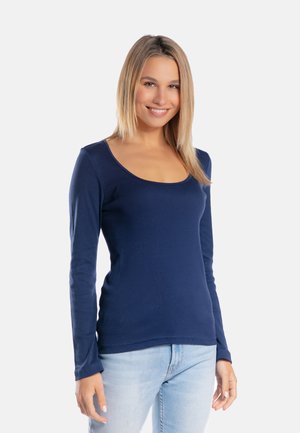 Młoda kobieta o prostych blond włosach, ubrana w granatową bluzkę z długim rękawem i jasnoniebieskie dżinsy, uśmiechająca się na tle białego, jednolitego tła.