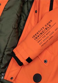 Veste isolante orange avec doublure verte, comportant du texte noir sur la manche, une fermeture éclair et un accent en forme de logo circulaire.