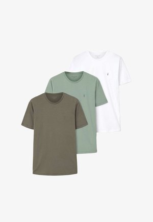 Młody mężczyzna stoi z rękami w kieszeniach, ubrany w oliwkowo zielony t-shirt z krótkim rękawem, czarne jeansy, czarny pasek i czarne buty na jasnym tle.