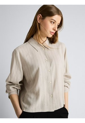 Femme aux longs cheveux bruns portant une chemise beige à fines rayures boutonnée avec les manches retroussées, regardant sur le côté, les mains dans les poches de son pantalon noir.