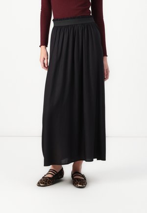 Femme portant une longue jupe noire, un haut côtelé bordeaux à manches longues et des chaussures plates imprimé léopard avec des brides noires, debout sur un fond blanc.
