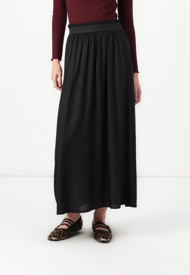 Femme portant une longue jupe noire, un haut côtelé bordeaux à manches longues et des chaussures plates imprimé léopard avec des brides noires, debout sur un fond blanc.