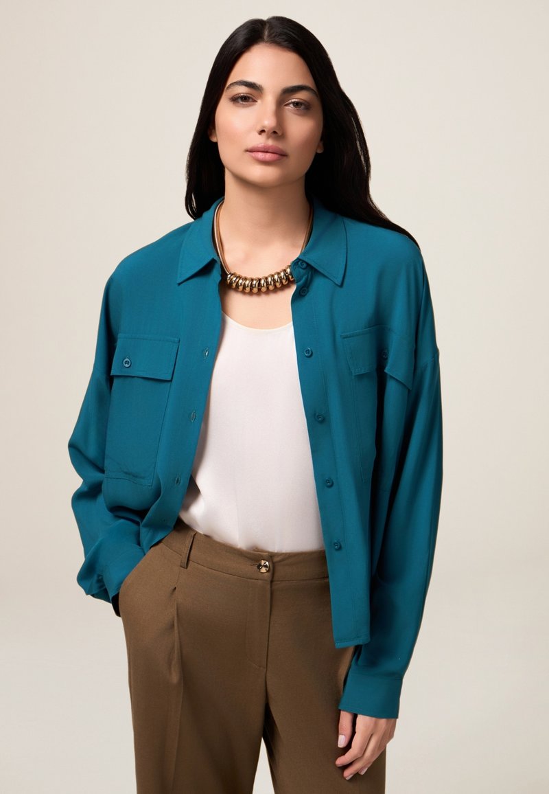Cămașă teal cu nasturi, cu două buzunare pe piept, purtată peste un top alb. Pantaloni maroon și un colier masiv din aur completează ținuta.