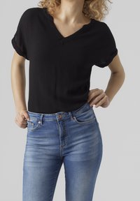 Schwarze, kurzärmelige Bluse mit V-Ausschnitt und strukturiertem Stoff. Kombiniert mit hellblauen Jeans aus Denim mit klassischer Passform und dezenten Ausbleichungen.