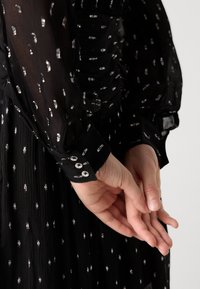 Abito nero con tessuto trasparente a pois argentati, dettaglio arricciato in vita e polsini con bottoni sulle maniche lunghe.