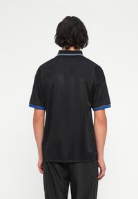 Άτομο με κοντά σκούρα μαλλιά που φοράει ένα μαύρο πουκάμισο πόλο με μανίκια σε μπλε απόχρωση και μαύρο παντελόνι, στέκεται με την πλάτη γυρισμένη σε έναν απλό φόντο.