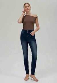 Egy nő barna vállra húzott felsőben és sötétkék, magas derékú, skinny farmernadrágban pózol, az egyik kezét a csípőjén tartja, a másikkal pedig a fülét érinti.