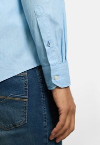 Hellblaues Langarmhemd mit strukturierter Oberfläche, geknöpftem Ärmelbündchen und leicht lockerer Passform, kombiniert mit dunkelblauen Jeans.