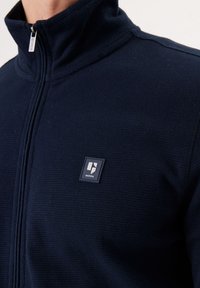 Giacca zippata blu navy con colletto alto, tessuto testurizzato e un'etichetta quadrata sul petto. Presenta una chiusura con zip e dettagli a costine.