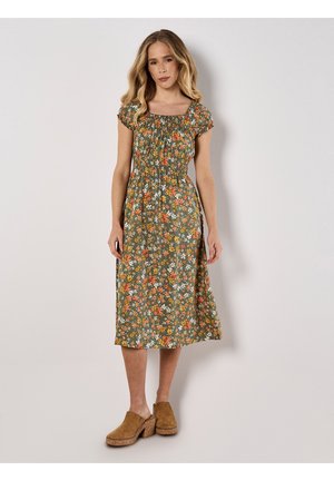 Blumenkleid in mittellanger Form in Grün mit orange und weißen Blumen. Hat kurze Puffärmel und einen gerafften Oberteil. Kombiniert mit hellbraunen Clogs.