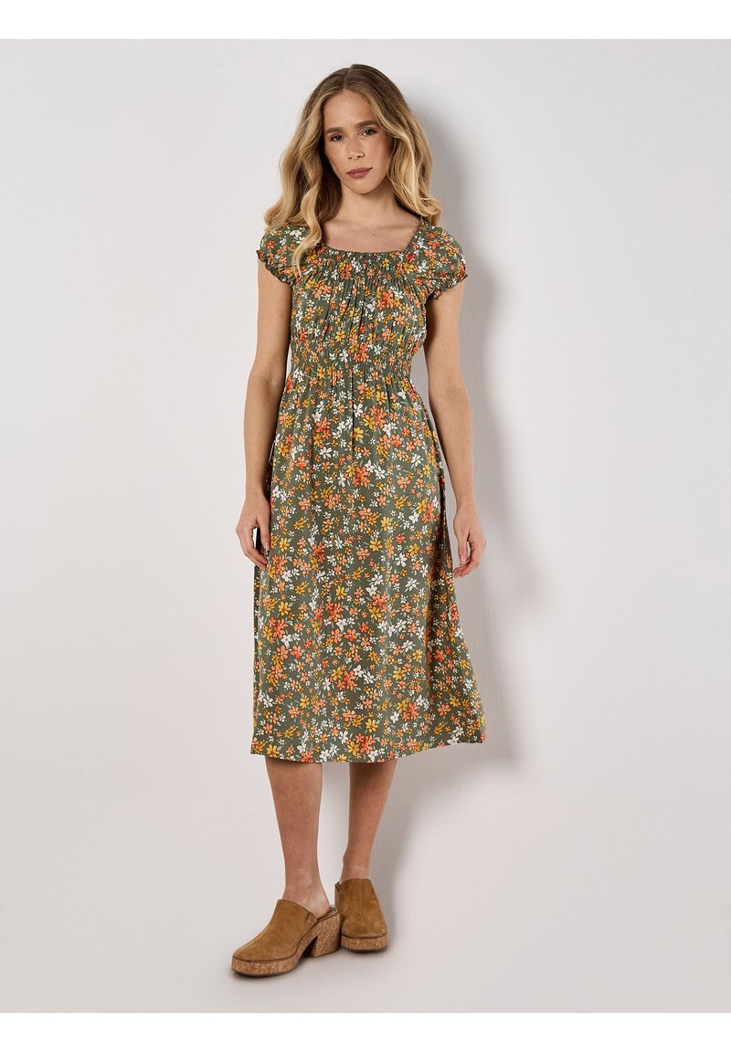 Blumenkleid in mittellanger Form in Grün mit orange und weißen Blumen. Hat kurze Puffärmel und einen gerafften Oberteil. Kombiniert mit hellbraunen Clogs.