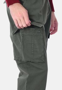 Pantaloni cargo verdi con tasca laterale, realizzati in un tessuto resistente. Presentano un dettaglio con bottone e cuciture a vista.