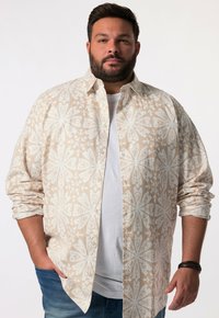 Camicia button-up color beige chiaro con un motivo floreale bianco, maniche lunghe arrotolate fino ai gomiti, indossata sopra una t-shirt bianca. Tessuto testurizzato.