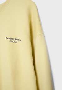 Svetle žlutá mikina s nápisem "Portobello Sundays LONDON" tištěným černě na prsou, zobrazující rameno a část rukávu na šedém pozadí.