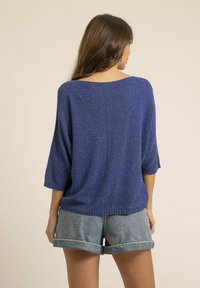 Pull bleu en maille avec des manches trois-quarts, présentant une coupe ample et une texture légèrement pailletée, associé à des shorts en denim.