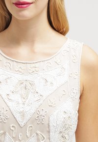 Top crème perlé avec broderies florales, présentant un décolleté élégant et une texture lisse. Les accents incluent des motifs de perles détaillés.