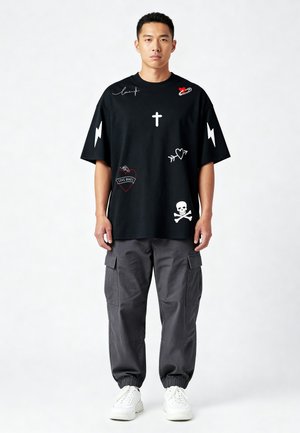 Homme debout portant un t-shirt noir oversize avec des symboles graphiques blancs et rouges, un pantalon cargo gris et des baskets blanches sur fond uni.