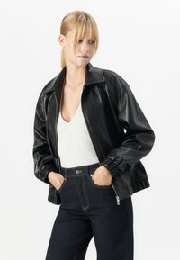 Kvinne med blondt hår i hestehale iført en svart skinnjakke, hvit V-hals topp og mørkeblå jeans med hendene i lommene.