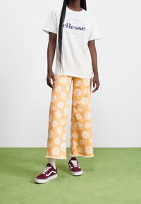 Weißes T-Shirt mit blauem und rotem Text, kombiniert mit gelben, floral gemusterten, weit geschnittenen Hosen und burgunderfarbenen Sneakers mit weißen Akzenten. Grüner Hintergrund.