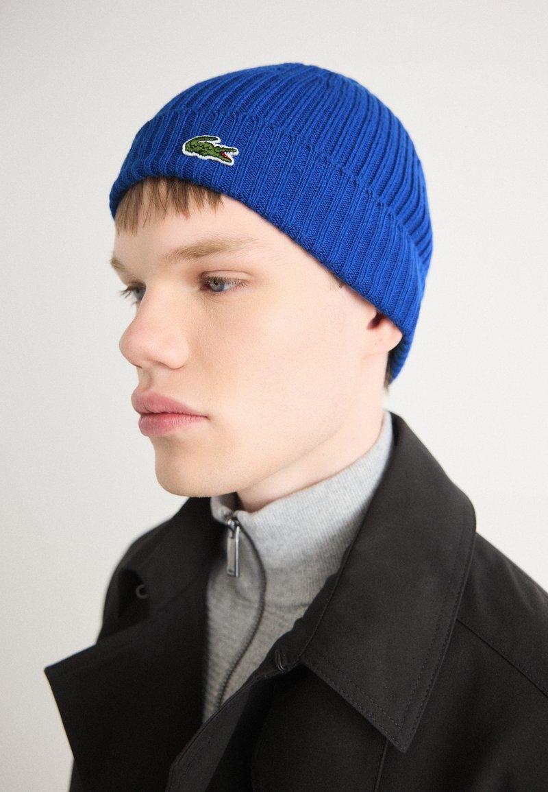 Blaue gerippte Strickmütze mit einem Krokodillogo, getragen von einem Model mit einem grauen Rollkragenpullover und einem schwarzen Mantel, der die Textur und Farbe hervorhebt.