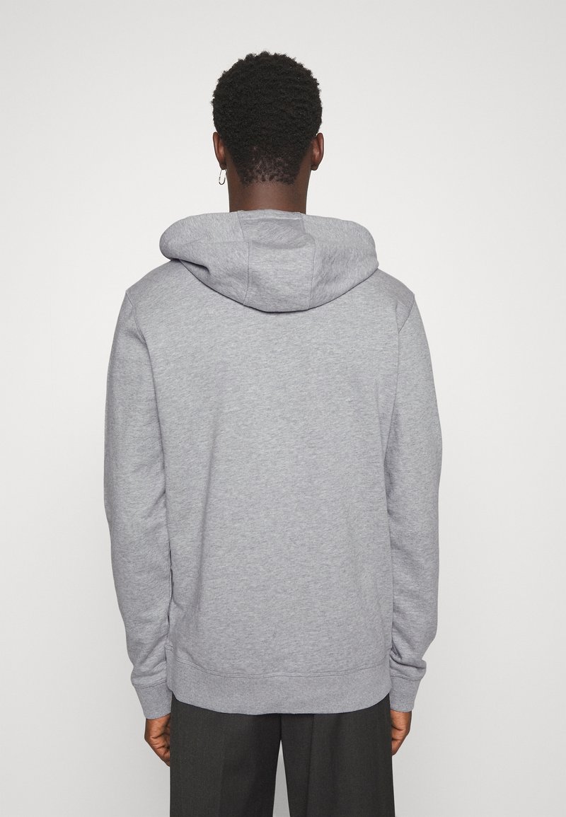 Homme aux cheveux bouclés foncés, debout de dos, portant un sweat à capuche gris clair uni et un pantalon foncé sur un fond blanc.