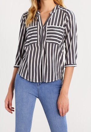Femme portant un chemisier rayé vertical noir et blanc avec des manches 3/4 et deux poches poitrine, associé à un jean bleu.